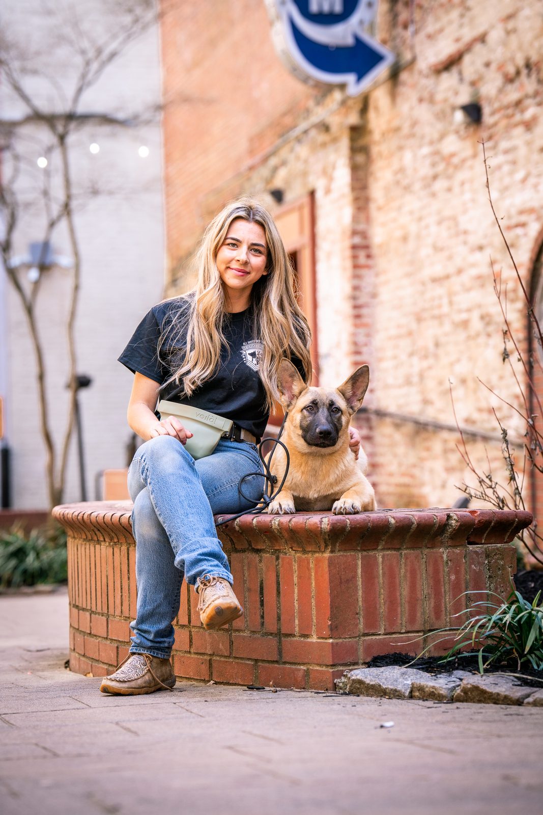 A trainer smiling beside a seated brown dog in a sunny setting